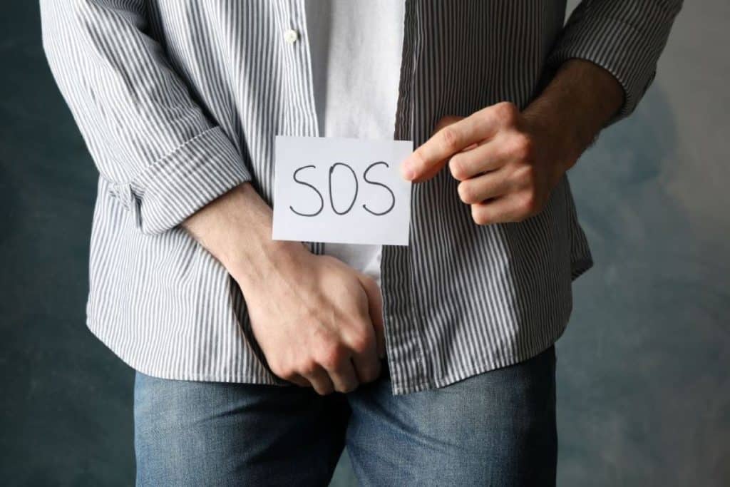 homem de camisa listrada e calça jeans segurando um cartão com a inscrição "SOS" próximo à sua região pélvica.