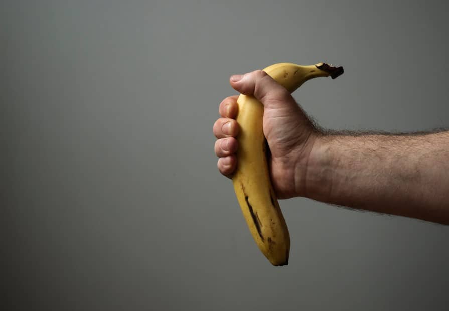 Homem segurando uma banana curvada em mãos como forma de representar a condição de pênis torto.