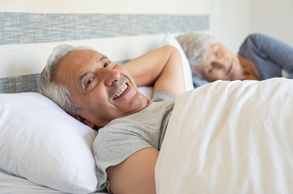 Homem, por volta dos 65 anos, deitado em sua cama ao lado da esposa, mulher por volta dos 60 anos, refletindo sobre até quantos anos o homem ejacula. 