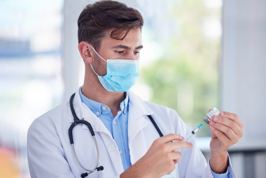 Médico está preparando uma injeção com grande foco e precisão. Ele segura uma seringa em uma mão e um frasco de medicamento na outra, sugerindo que está prestes a proceder com uma aplicação ou um tratamento. Este tipo de procedimento pode estar associado a tratamentos médicos inovadores como os destinados a engrossar o pênis.