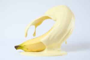 A imagem apresenta uma composição artística onde uma banana amarela está parcialmente coberta por um líquido viscoso de cor creme claro, criando uma representação visual que pode evocar temas como a cor do esperma masculino.