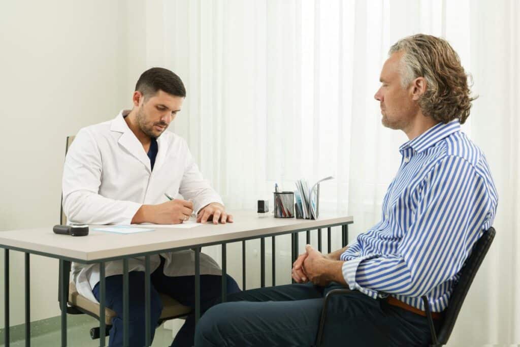 Prótese peniana maleável: o que é e como funciona? consulta médica, onde um médico jovem e atento está sentado à mesa, escrevendo em um papel enquanto conversa com seu paciente sobre protese peniana maleavel