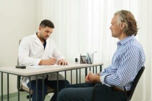 consulta médica, onde um médico jovem e atento está sentado à mesa, escrevendo em um papel enquanto conversa com seu paciente sobre protese peniana maleavel