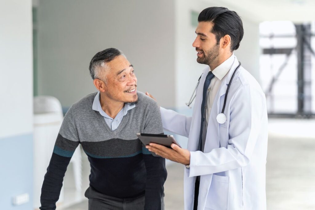 Prótese peniana semirrígida: como funciona, indicações e benefícios do implante médico, usando um jaleco branco e segurando um tablet, parece estar explicando algo ao paciente, que sorri enquanto olha para ele, proporcionando uma interação positiva. Este cenário pode representar uma discussão sobre opções de tratamento para disfunção erétil, incluindo a "prótese peniana semirrígida".