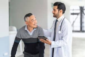médico, usando um jaleco branco e segurando um tablet, parece estar explicando algo ao paciente, que sorri enquanto olha para ele, proporcionando uma interação positiva. Este cenário pode representar uma discussão sobre opções de tratamento para disfunção erétil, incluindo a "prótese peniana semirrígida".