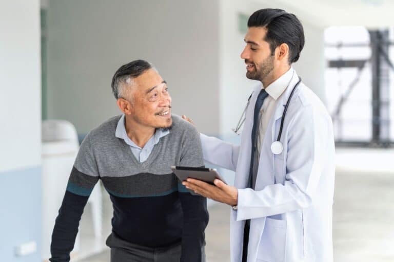 médico, usando um jaleco branco e segurando um tablet, parece estar explicando algo ao paciente, que sorri enquanto olha para ele, proporcionando uma interação positiva. Este cenário pode representar uma discussão sobre opções de tratamento para disfunção erétil, incluindo a "prótese peniana semirrígida".